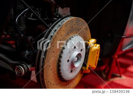 A close up shows a carbon ceramic disc, vented rotor, and gold caliper on a luxury sports SUV or coupe in a pro workshop, wheel off, parts lit with bright directional light. 138430136