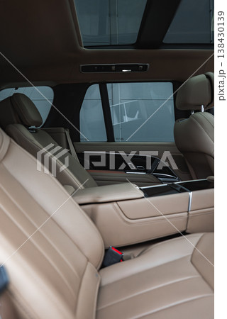 Close up of a luxury SUV rear cabin shows tan perforated seats, wide armrests, glossy black console, tinted windows, and a panoramic glass roof indoors in a showroom. Close up of a luxury SUV rear cabin shows tan perforated seats, wide armrests, glossy black console, tinted windows, and a panoramic glass roof indoors in a showroom. 138430139