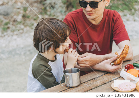 Close-up view of father hugs his school boy son on a family picnic. Child kid and his dad taking a rest and enjoying a picnic while hiking. Boy smiles and bites the bread donut on a picnic 138431996