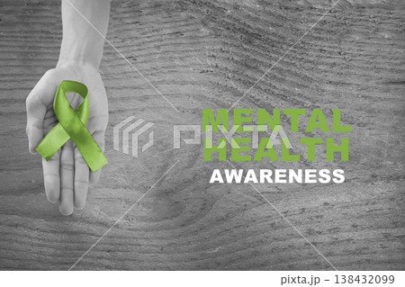 Grayscale image of a hand holding a green ribbon with Mental Health Awareness text on a rustic wooden background. A powerful symbol of support for psychological well-being Grayscale image of a hand holding a green ribbon with Mental Health Awareness text on a rustic wooden background. A powerful symbol of support for psychological well-being 138432099