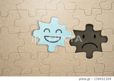 A light blue puzzle piece with a happy smile sits next to an empty space showing a sad face, symbolizing the contrast between happiness and sadness in mental health and emotions 138432104