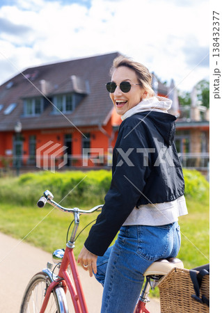 Smiling woman riding bicycle on sunny day near colorful house green park landscape. Active lifestyle moment shows freedom, happiness and outdoor recreation relaxed urban cycling summer atmosphere Smiling woman riding bicycle on sunny day near colorful house green park landscape. Active lifestyle moment shows freedom, happiness and outdoor recreation relaxed urban cycling summer atmosphere 138432377