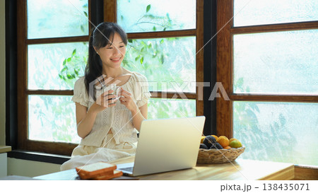 Young woman drinks coffee while using laptop at kitchen counter. 138435071