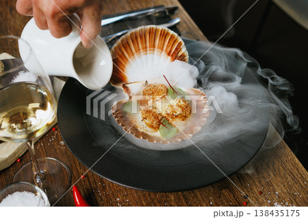 Grilled scallops served in a shell with avocado mousse and microgreens. Elegant seafood dish presented with dry ice smoke. Gourmet cuisine in a fine dining restaurant Grilled scallops served in a shell with avocado mousse and microgreens. Elegant seafood dish presented with dry ice smoke. Gourmet cuisine in a fine dining restaurant 138435175