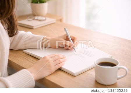 コーヒーを飲みながらノートに文字を書く女性の手元 138435225