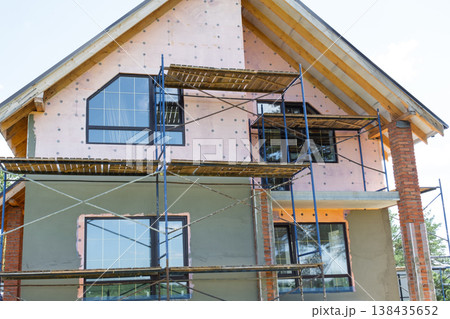 House under facade plastering, exterior scaffolding for render application, insulation layer, surface leveling, construction wall finish, decorative coating. 138435652