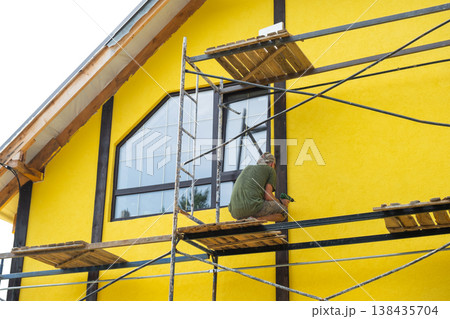 Applying finish paint to facade plaster, house exterior wall, renovation project, yellow color, wood decorative strips, maintenance step, final wall protection. Applying finish paint to facade plaster, house exterior wall, renovation project, yellow color, wood decorative strips, maintenance step, final wall protection. 138435704