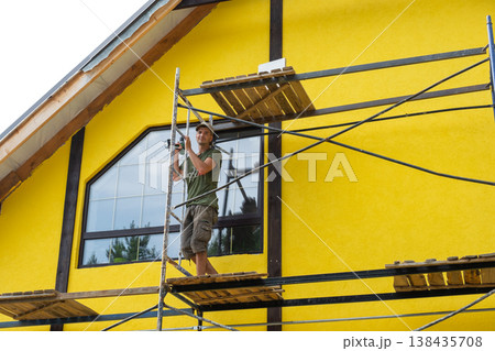 House painting at exterior stage, finishing yellow facade color, plaster coated wall, worker on scaffold, possible timberboard decor, renovation completion step. 138435708