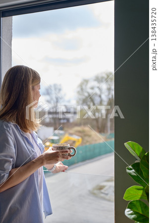 Woman holding mug and looking through window in daylight. Concept of coffee break Woman holding mug and looking through window in daylight. Concept of coffee break 138435720