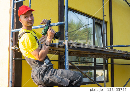 Builder in red cap with screwdriver on scaffolding, house facade, home repair, instrument kit, handyman visiting for at-home craft, maintenance and improvement. 138435735