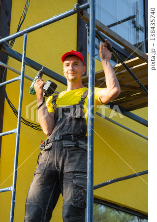 Repairman in overalls, on scaffold with paint roller, facade painting, consulting for exterior house renovation, on-site service, handyman tool kit. Repairman in overalls, on scaffold with paint roller, facade painting, consulting for exterior house renovation, on-site service, handyman tool kit. 138435740