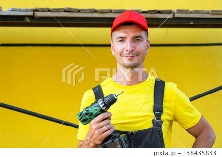 Builder with cordless drill and toolkit, standing at house under construction, at-home repairs, professional contractor business, DIY improvement skills. Builder with cordless drill and toolkit, standing at house under construction, at-home repairs, professional contractor business, DIY improvement skills. 138435833