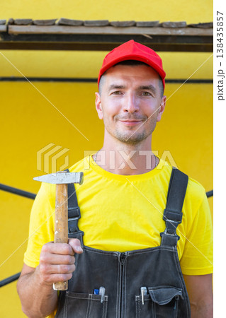 Portrait of builder in red cap holding hammer, private handyman business, home repairs, carpentry, on-site DIY construction, home building expert. Portrait of builder in red cap holding hammer, private handyman business, home repairs, carpentry, on-site DIY construction, home building expert. 138435857