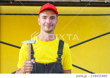 Young handyman in red cap holding hammer, home construction, carpentry, nailing, repair work, private business, DIY services, on-site call, craftsmanship. Young handyman in red cap holding hammer, home construction, carpentry, nailing, repair work, private business, DIY services, on-site call, craftsmanship. 138435860