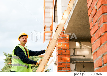 Builder secures roof beam installation, construction site safety, masonry wall, frame support, teamwork, project progress, structure stabilization, project oversight. Builder secures roof beam installation, construction site safety, masonry wall, frame support, teamwork, project progress, structure stabilization, project oversight. 138436026
