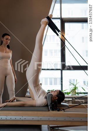 Woman holding legs vertical with help on pilates reformer for flexibility training by bright window. Flexibility training, pilates reformer, vertical legs, bright window, assistance. 138437687