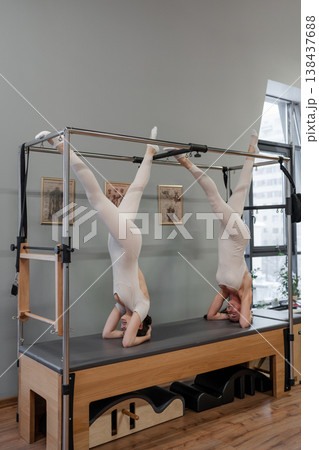 Pilates duo balancing together in upside-down leg pose on reformer trainers in gray exercise studio. Upside-down, duo, pilates, reformer, leg pose. 138437688