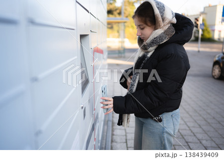 Girl uses a package delivery box on a city street during the day, scanning qr code to open parcel locker 138439409