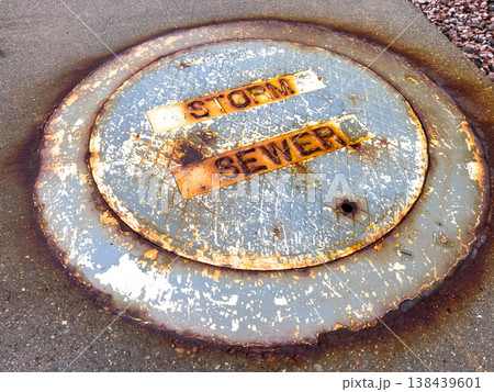 A close-up of a corroded manhole cover stamped with STORM SEWER. The rust and oxidation form an abstract texture around the edges. A close-up of a corroded manhole cover stamped with STORM SEWER. The rust and oxidation form an abstract texture around the edges. 138439601