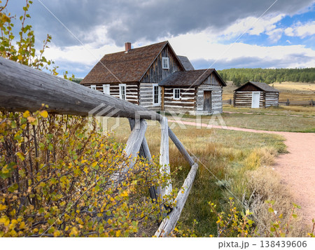 A preserved log homestead stands behind a rustic wooden fence surrounded by golden autumn foliage in Florissant, Colorado. The bright daylight and open landscape highlight the rural charm and 138439606