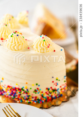 A vertical shot of a vanilla birthday cake slice with bright sprinkles and frosting on a white plate. The image has a soft, celebratory aesthetic with pastel lighting. 138439636