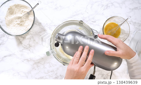 Hands operate a stand mixer blending cookie dough mixture inside a glass bowl on a marble surface. Flour and beaten eggs are arranged nearby, emphasizing organized baking preparation. Bright lighting Hands operate a stand mixer blending cookie dough mixture inside a glass bowl on a marble surface. Flour and beaten eggs are arranged nearby, emphasizing organized baking preparation. Bright lighting 138439639
