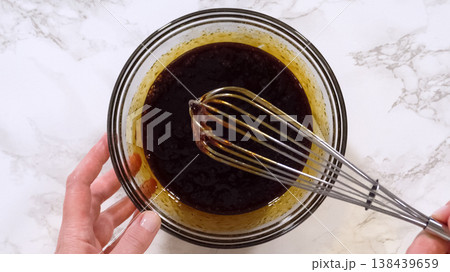 Hands whisk a dark liquid mixture in a glass bowl placed on a marble surface. The mixture has a glossy texture, representing the early stage of cookie dough preparation. Bright lighting highlights the Hands whisk a dark liquid mixture in a glass bowl placed on a marble surface. The mixture has a glossy texture, representing the early stage of cookie dough preparation. Bright lighting highlights the 138439659