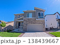 A two-story modern suburban home with a stone accent facade and large garage under clear blue sky. The house is surrounded by neighboring homes and landscaped with grass and a small tree. Captured 138439667