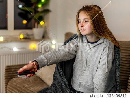 Little girl sits on sofa wrapped in blanket holding remote and watching TV at home in evening 138439811
