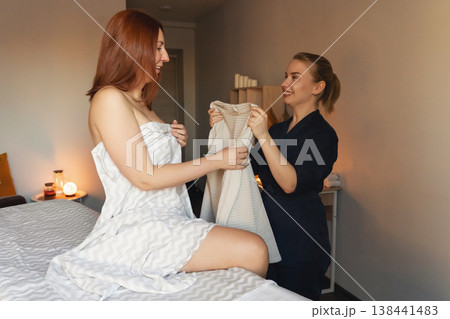 Woman wrapped in a towel sitting on a massage table, receiving a bathrobe from a smiling therapist at a wellness center Woman wrapped in a towel sitting on a massage table, receiving a bathrobe from a smiling therapist at a wellness center 138441483