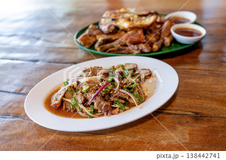 Thai spicy grilled pork salad known as Nam Tok Moo on a white plate. In the background is grilled chicken with dipping sauce on a wooden table. Authentic Northeastern Thai cuisine. 138442741