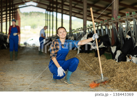 Portrait of woman working on livestock farm posing in cowshed near stall with herd of cows 138443369