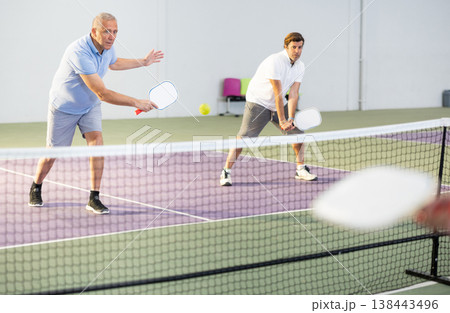 Senior man playing doubles pickleball with male partner indoors Senior man playing doubles pickleball with male partner indoors 138443496