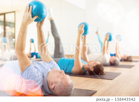 Class of male and women of different ages in active wear pressing balls in hands exercise during Pilates training together in gym Class of male and women of different ages in active wear pressing balls in hands exercise during Pilates training together in gym 138443542