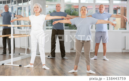 Experienced mature female ballet dancer, dressed in sportswear executing dance moves with focused precision among group of aged people rehearsing in ballet studio Experienced mature female ballet dancer, dressed in sportswear executing dance moves with focused precision among group of aged people rehearsing in ballet studio 138443759