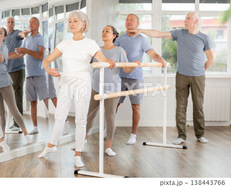 Group of elderly dancers engaged in ballet in dance studio perfoms exercise near ballet barre, senior male choreographer helps to coordinate movements correctly Group of elderly dancers engaged in ballet in dance studio perfoms exercise near ballet barre, senior male choreographer helps to coordinate movements correctly 138443766