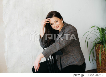 A beautiful smiling brunette poses while sitting on a chair. Businesswoman. Plus-size model. Happy woman concept.  138450752