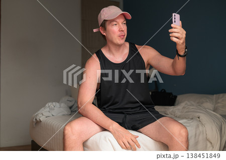 Caucasian Man in Pink Cap Taking Selfie with Smartphone on Bed in Bedroom Caucasian Man in Pink Cap Taking Selfie with Smartphone on Bed in Bedroom 138451849