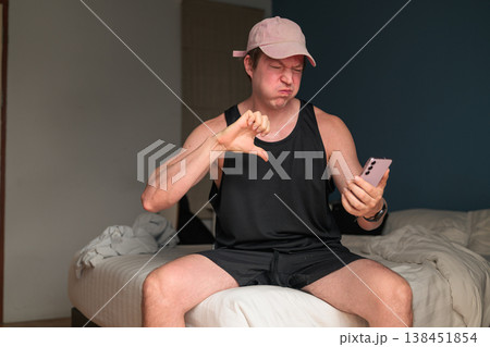 Caucasian Man in Pink Cap Giving Thumbs Down to Smartphone on Bed in Bedroom Caucasian Man in Pink Cap Giving Thumbs Down to Smartphone on Bed in Bedroom 138451854