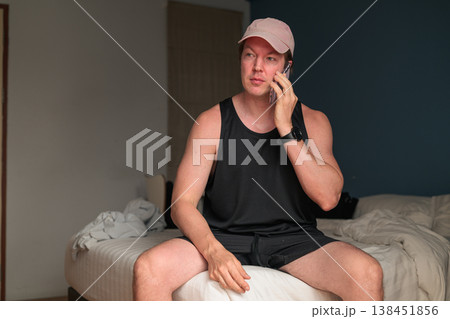 Caucasian Man in Pink Cap Talking on Smartphone on Bed in Bedroom Caucasian Man in Pink Cap Talking on Smartphone on Bed in Bedroom 138451856