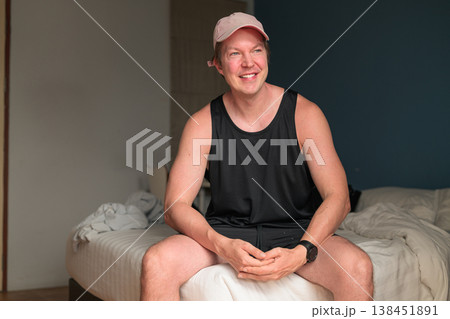 Smiling Thinking Man in Pink Cap Sitting on Bed in Minimalist Bedroom 138451891