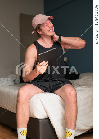 Caucasian Man with Neck Pain Holding Tablet on Bed in Minimalist Bedroom 138451936