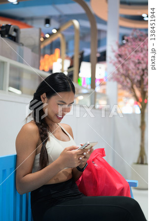 Asian Woman in Fitness Wear Using Smartphone on Bench in Mall with Copy Space 138451944