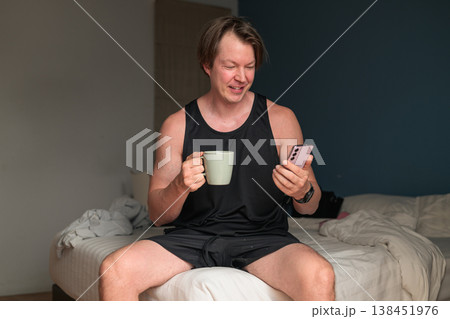 Smiling Caucasian Man in Tank Top Using Smartphone and Holding Coffee Mug on Bed 138451976