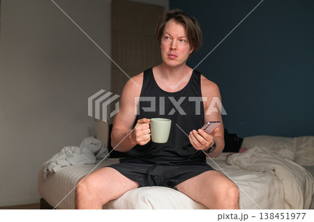 Mid Adult Caucasian Man Sitting on Bed with Coffee and Smartphone in Minimalist Bedroom 138451977