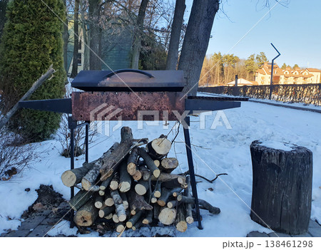 Winter, in the yard with a metal barbecue with a lid and stacked firewood under it. Snow, bare trees, an atmosphere of comfort and preparation for cooking outdoors. Countryside vacation, barbecue 138461298