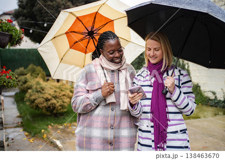 Diverse women friends smiling using smartphone under umbrellas in rain Diverse women friends smiling using smartphone under umbrellas in rain 138463670