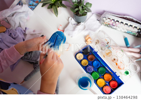 A childs hands paint a dolls hair blue with a brush on a table with a paint cup and palette surrounded by plants and art supplies creative hobby crafting diy concept 138464298