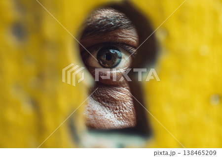 Eye peers through keyhole in yellow surface, conveying mystery and observation with intense focus, bringing an element of intrigue. Eye peers through keyhole in yellow surface, conveying mystery and observation with intense focus, bringing an element of intrigue. 138465209