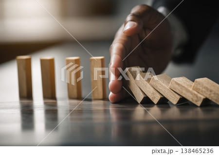 Preventing a chain reaction is shown with a hand stopping falling wooden blocks, representing stability and risk management. 138465236
